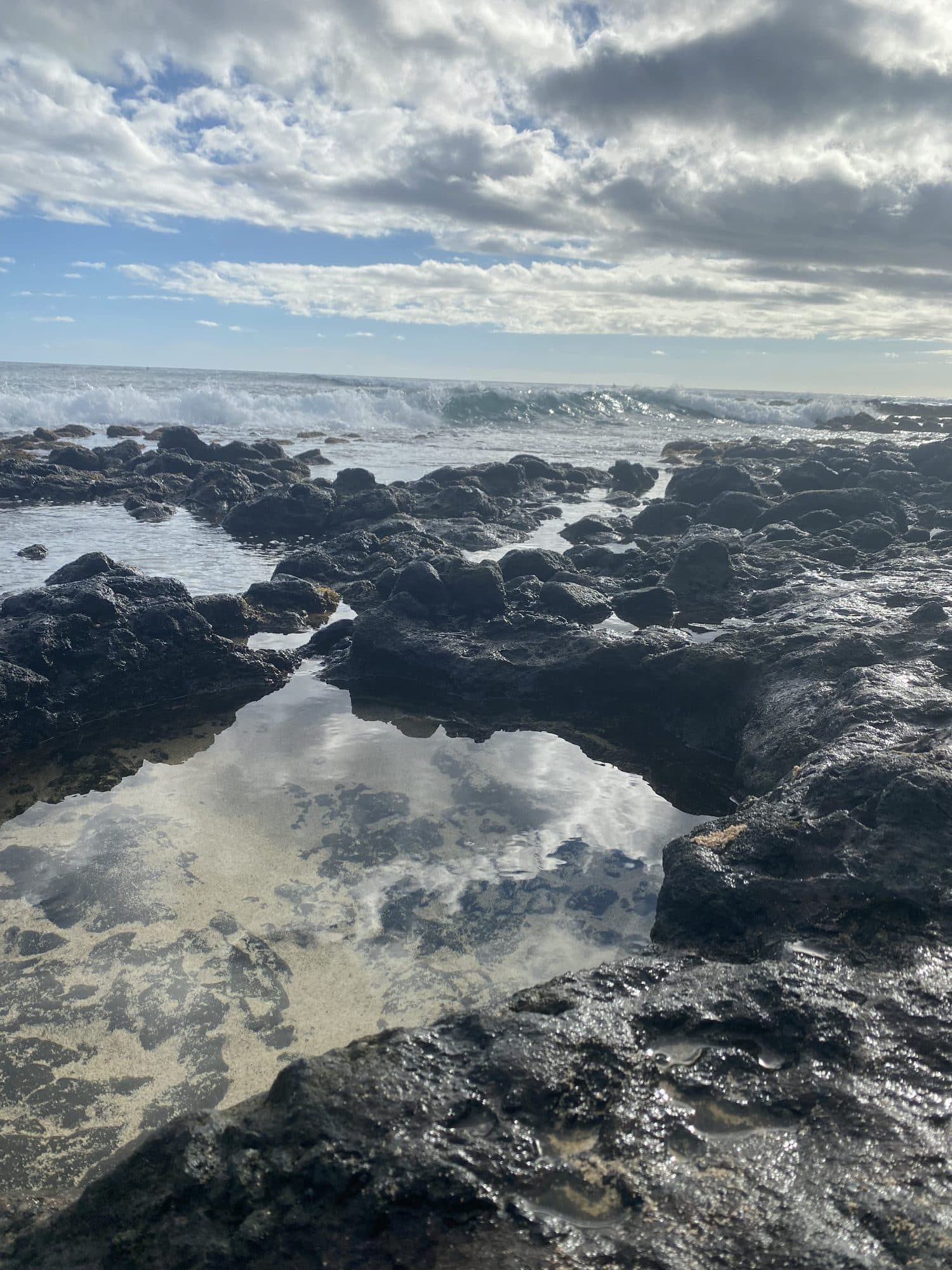 A tide pool among dark rocks holding the sky and clouds