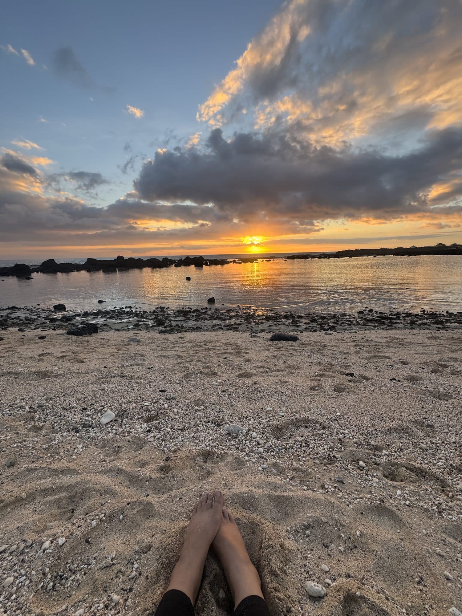 Bare feet resting in the sand as the sun sets over the water