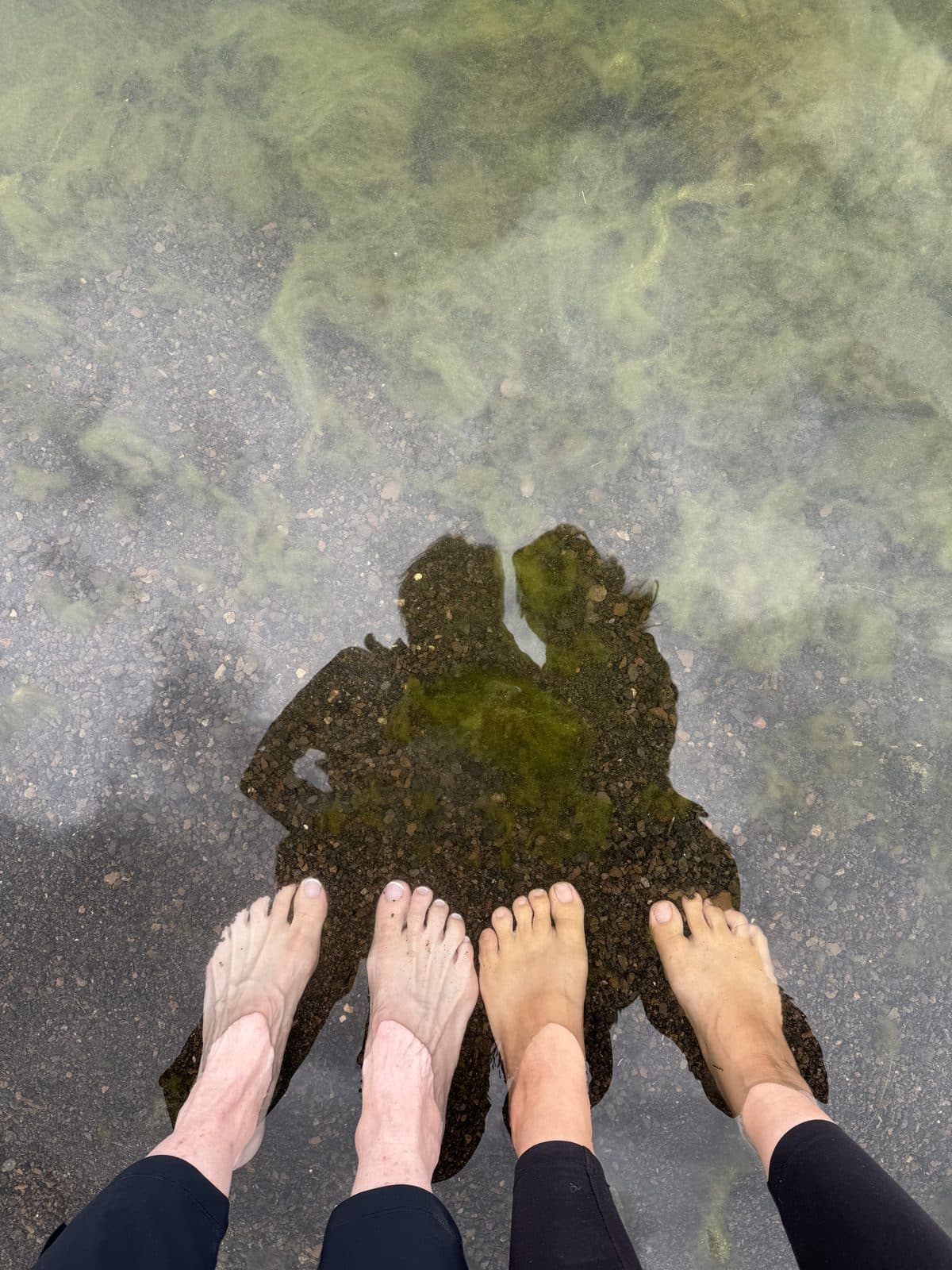 Two pairs of bare feet in clear shallow water, their shadows leaning together